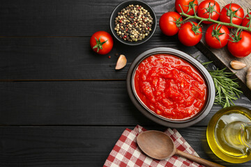 Homemade tomato sauce in bowl, spoon and fresh ingredients on black wooden table, flat lay. Space for text