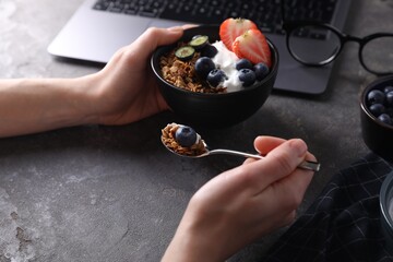 Woman eating tasty granola with yogurt and berries at workplace, closeup