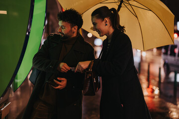 A couple stands under an umbrella on a rainy day while a man withdraws money from an ATM machine.