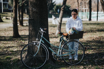 A young adult female dressed in casual clothing stands by a classic bicycle with a basket, enjoying a peaceful day in a sunny park.