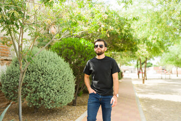 Young hispanic man in sunglasses and black t-shirt posing for a mockup, with a natural park background and casual posture