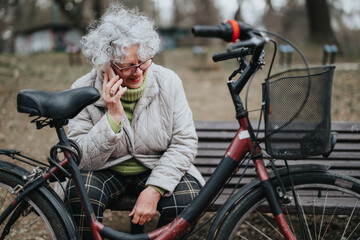 Mature woman with glasses and white hair talking on the phone while resting with her bicycle in an...