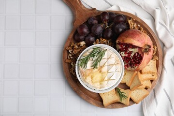 Board with tasty baked camembert, croutons, grapes, walnuts and pomegranate on white tiled table, top view. Space for text