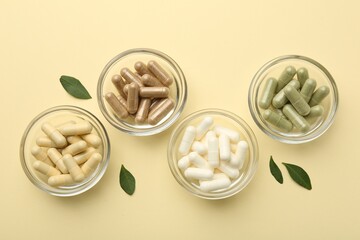 Different vitamin capsules in bowls and leaves on pale yellow background, flat lay