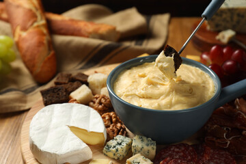 Dipping piece of bread into fondue pot with melted cheese at wooden table, closeup