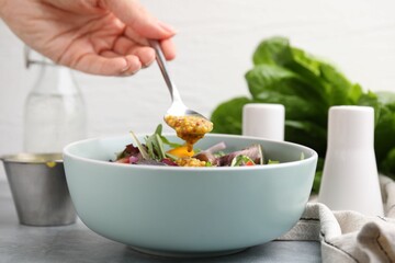 Woman pouring tasty vinegar based sauce (Vinaigrette) from spoon into bowl with salad at grey...