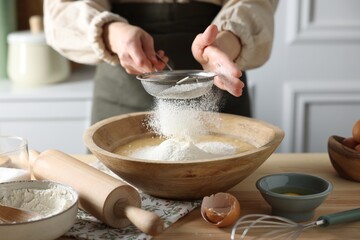 Making dough. Woman sifting flour into bowl at wooden table in kitchen, closeup