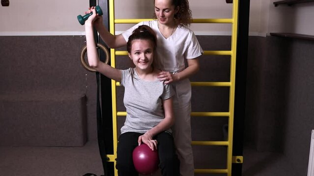 Physician Helps Happy Disabled Child Raise Dumbbell, Do Physical Exercises In Rehabilitation Room. Kid With Special Needs In Gym. Rehabilitation. Horizontal plane. Slow Motion