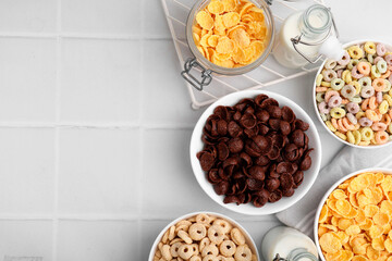 Different delicious breakfast cereals and milk on white tiled table, flat lay. Space for text