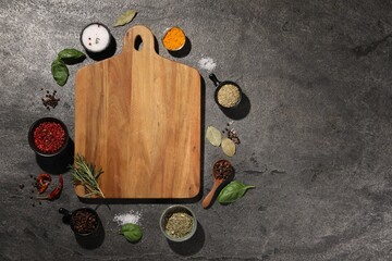 Wooden cutting board surrounded by spices on gray textured table, flat lay. Space for text