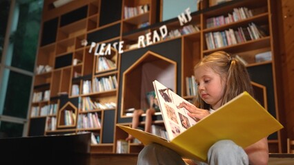 Young smart caucasian girl picking reading a book while sitting at library. Clever child learning,...