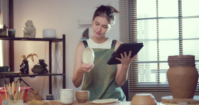 Focused female potter examining a ceramic plate while cross-referencing designs on a digital tablet in her studio.
