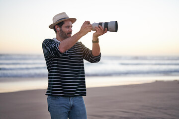 mature man photographer holding a pro photo camera taking pictures with zoom lens on the beach at...