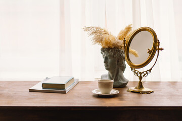 Gold-framed mirror stands on a wooden table, beside a classical bust vase adorned with pampas...
