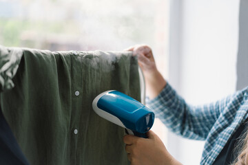Woman Using a Blue Handheld Steamer on a Green Garment at Home During Daytime. Natural daylight...