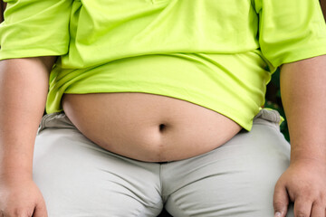 A fat man wears a yellow shirt and sits in a park with a bloated stomach. There is a lot of excess...