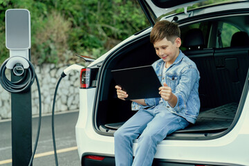 Little boy sitting on car trunk, using tablet while recharging eco-friendly car from EV charging...