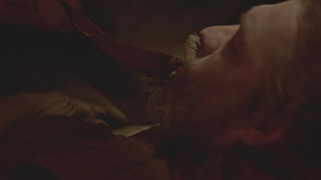 Close up side view of female hands giving a decoction to a sick man. Media. Bearded man drinking from a wooden spoon in dark room.