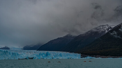 A wall of cracked blue ice rises above a glacial lake. Canoes with tourists sail through the turquoise water to the glacier. Coastal mountains in clouds and fog. Perito Moreno. El Calafate. Argentina