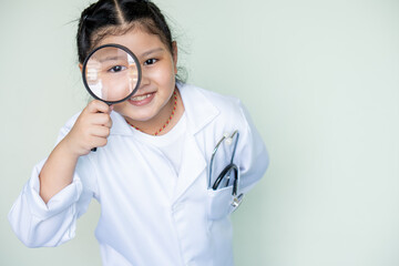 Cute girl asian in a medical play a doctor  .magnifying glass and portrait of a scientist girl in room with eye for scientific research. Face of a  kid student with magnifier for medical science.