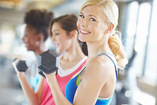 Portrait, dumbbell and happy woman in gym for exercise, training or fitness with friends. Face, smile and strong girl weightlifting for workout, wellness or muscle for healthy body at sports club - Powered by Adobe