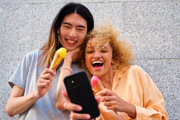 Two multiracial happy friends with ice creams taking a selfie in summer at street.