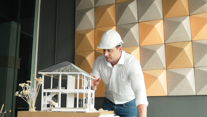 Closeup portrait of caucasian male architect engineer inspect and check house model construction with project plan and architectural model placed on table. Business design concept. Manipulator.