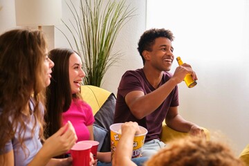 Group of friends laughing, drinking and eating popcorn while watch a movie at shared flat.
