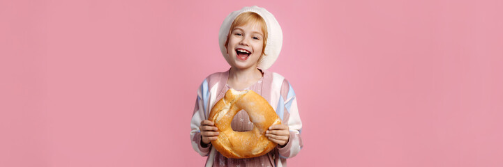 A Caucasian child in a white chef hat and striped apron holds a large golden bagel, pink...