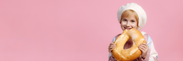 A Caucasian child in a white chef hat and striped apron holds a large golden bagel, pink...