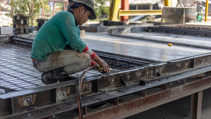 Worker with safety hat is welding pieces of metal mold together for precast floor production. Technician is preparing a block for concrete floor making.