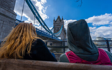 A lesbian couple sitting on a wooden bench on the River Thames promenade with the Tower Bridge in...