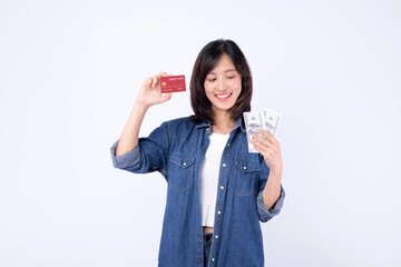 Asian woman wearing denim jean is holding dollar and credit card against a white background,...