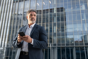 Focused serious latin middle age male entrepreneur businessman holding mobile cell phone, looking aside. Mature hispanic senior business man using smartphone cellphone at office building. Copy space