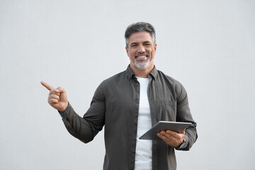 Hispanic mature adult professional business man holding touchscreen pc computer, looking at camera, pointing aside. Smiling Indian businessman CEO using tablet application, isolated white background