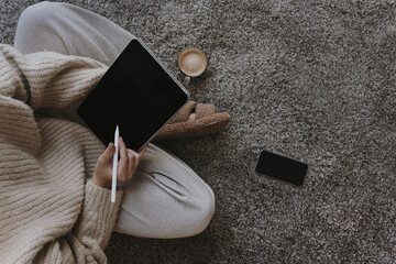 Top view tablet mockup with copy space. Woman wearing beige clothes sitting on carpet with coffee cup and works using digital tablet. Cozy work at home concept