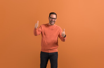 Portrait of ecstatic male professional in eyeglasses gesturing thumbs up sign on orange background
