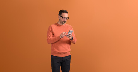 Young handsome entrepreneur in eyeglasses messaging over mobile phone on isolated orange background