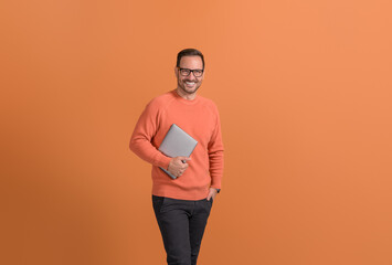 Positive young businessman with hand in pocket holding wireless computer against orange background