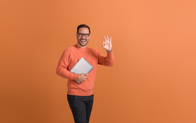 Successful happy businessman holding laptop and showing OK gesture over isolated orange background