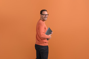 Side view of happy young businessman holding laptop and looking over shoulder on orange background