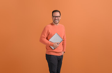 Portrait of young inspired businessman holding wireless computer and smiling on orange background