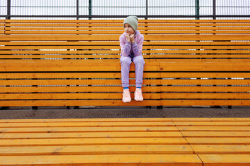 A girl sits on a wooden bench in a sports tribune with her hands on her knees. Child athlete in a...
