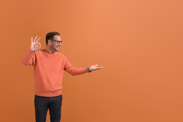 Happy young salesman showing OK sign and looking at empty palm while standing on orange background