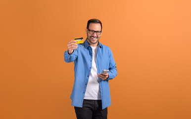 Portrait of happy young man holding mobile phone and advertising credit card on orange background