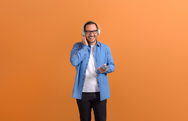 Portrait of handsome man listening music over headphones and using smartphone on orange background