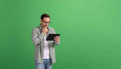 Thoughtful businessman with hand on chin analyzing strategy over digital tablet on green background