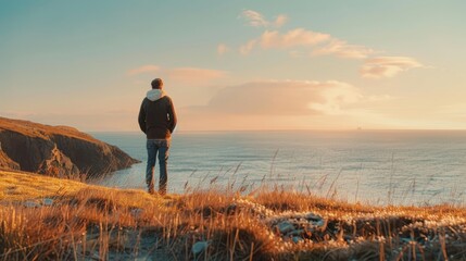 Man in a hoodie and jeans stands contemplatively overlooking the ocean from a cliff during a sunset, evoking a sense of solitude and reflection