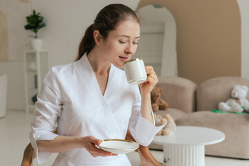 Medicine, healthcare and people concept - Friendly female gp physician doctor in white uniform drinking coffee tea having breakfast while working in the clinic office. Part of a series