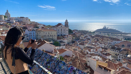 A tourist photographs an aerial view of the Alfama district with Tagus river in the background.